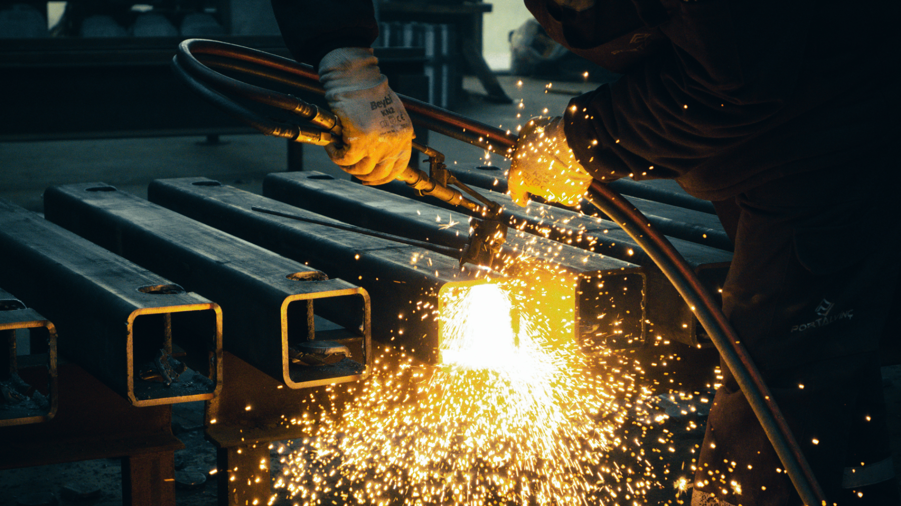 Welder using a torch to cut heavy steel box sections in the workshop.